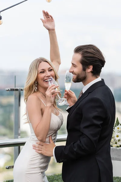 Cheerful blonde bride holding champagne near groom on terrace — Photo de stock