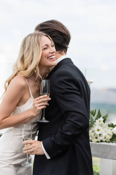 Весела наречена тримає келих шампанського біля елегантного нареченого в костюмі на терасі — стокове фото