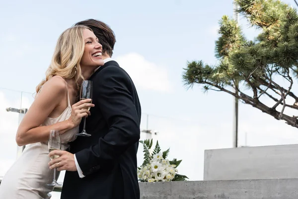 Smiling bride holding champagne and standing near groom and bouquet on terrace - foto de stock