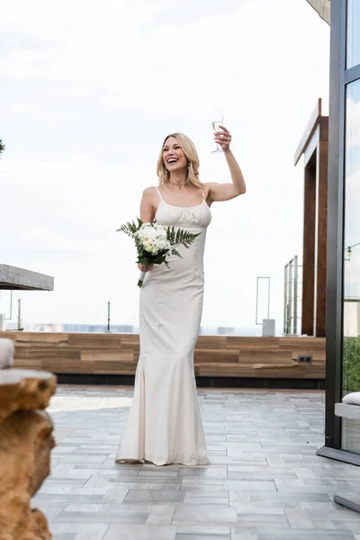 Positive bride holding glass of champagne and bouquet on terrace of restaurant — Stock Photo