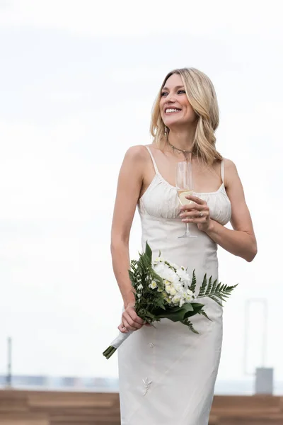 Blonde bride in wedding dress holding flowers and champagne outdoors — Foto stock