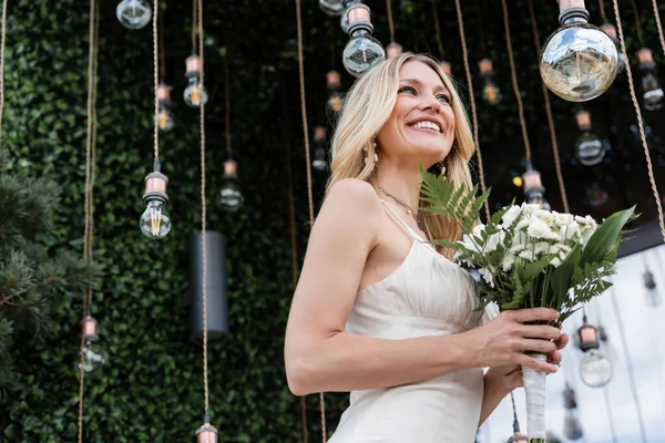 Vista a basso angolo della sposa bionda che tiene bouquet da sposa vicino alle lampadine sulla terrazza — Foto stock