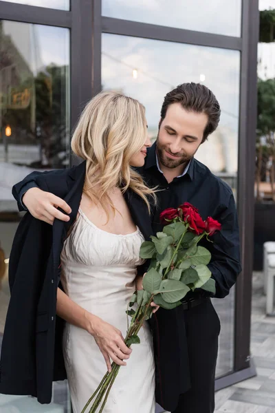 Brunette man wearing jacket on girlfriend with red roses on terrace - foto de stock