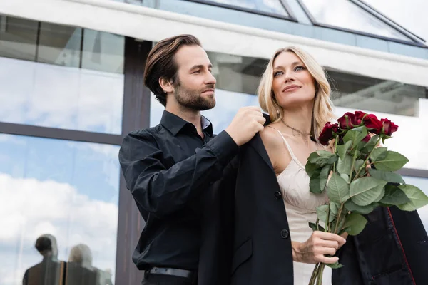 Low angle view of young man wearing jacket on blonde girlfriend with roses during date outdoors — Stock Photo
