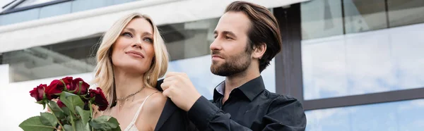 Bearded man wearing jacket on girlfriend with red flowers outdoors, banner — Stockfoto