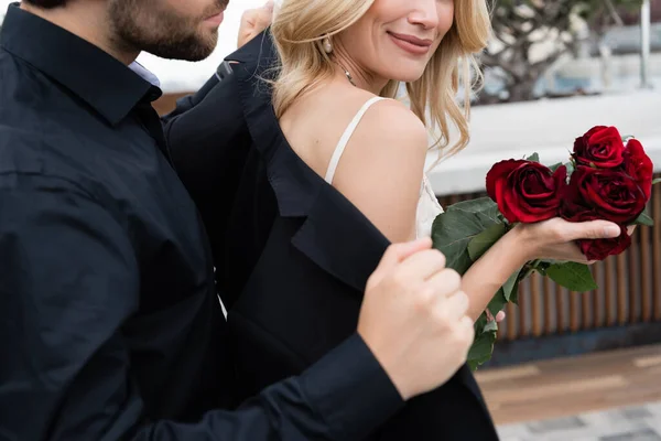 Cropped view of man wearing jacket on blonde girlfriend with roses outdoors — Stockfoto