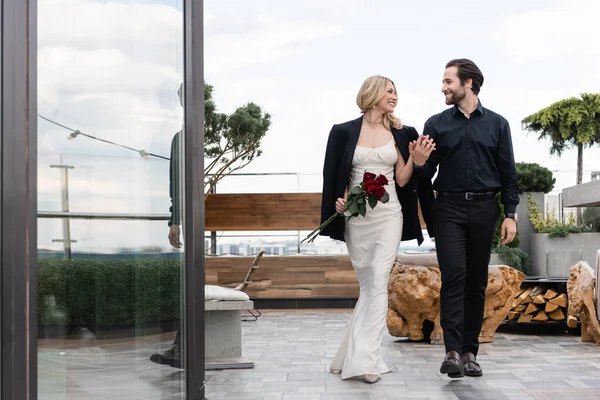 Side view of smiling woman in dress and jacket holding roses and hand of boyfriend on terrace of restaurant - foto de stock