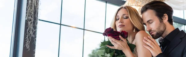 Brunette man in suit touching shoulder of blonde girlfriend with roses in restaurant, banner — Stockfoto