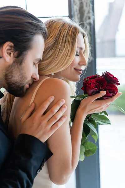 Side view of brunette man kissing smiling girlfriend with roses in restaurant — Photo de stock