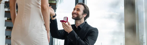 Smiling man in suit holding engagement ring near girlfriend in restaurant, banner — Stockfoto