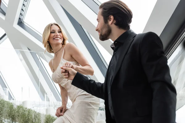 Low angle view of smiling woman in dress holding hand of blurred boyfriend in restaurant - foto de stock