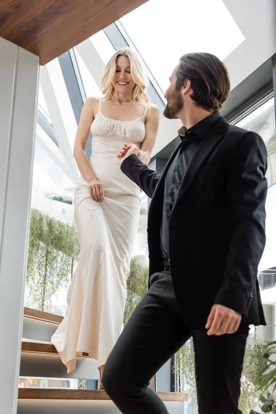 Elegant man in suit holding hand of girlfriend in dress on stairs in restaurant - foto de stock