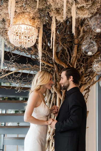 Side view of smiling elegant couple holding hands near decorative tree in restaurant — Fotografia de Stock