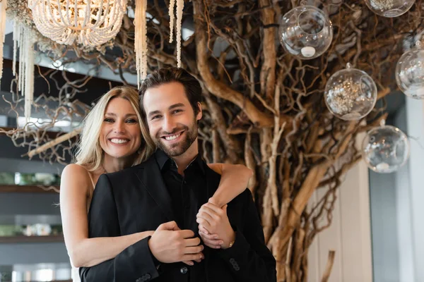 Smiling woman hugging boyfriend in suit and looking at camera in restaurant — Stock Photo