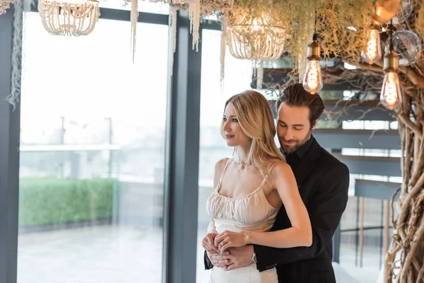 Elegant man hugging blonde girlfriend under light bulbs in restaurant — Foto stock