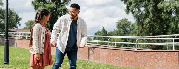 Cheerful asian dad holding hand of kid and talking in park, banner - foto de stock