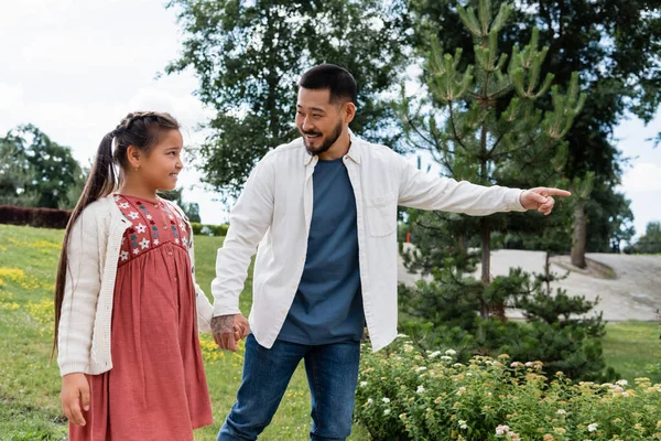 Cheerful asian father pointing with finger near daughter in park - foto de stock