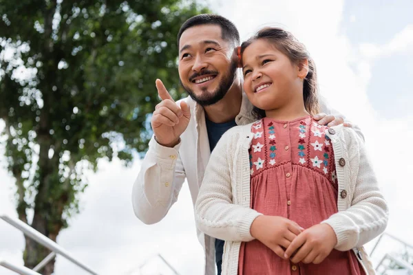 Low angle view of smiling asian father pointing with finger near daughter in park — Stock Photo