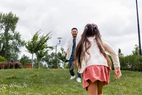 Smiling asian man looking at running daughter in summer park — Photo de stock