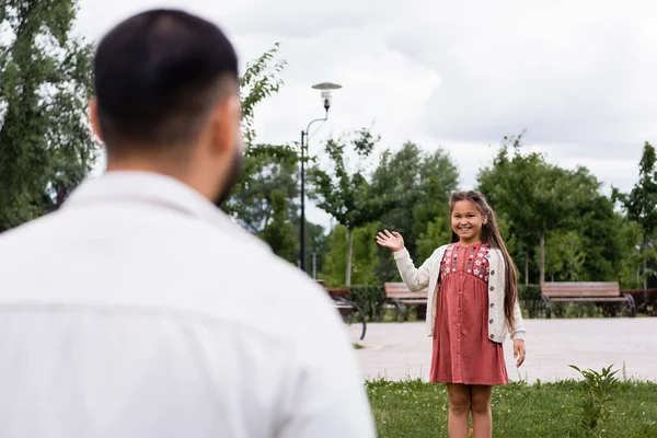 Smiling asian girl waving hand at blurred dad in summer park — Stockfoto