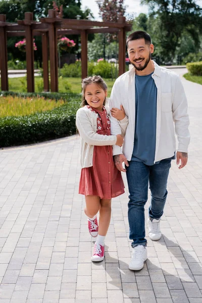 Позитивна азіатська дівчина тримає руку тата під час прогулянки в літньому парку — стокове фото