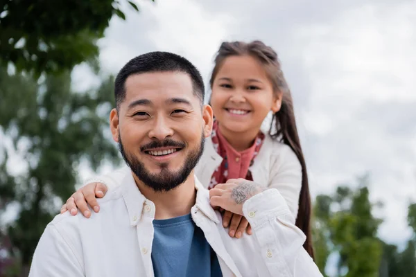 Smiling asian man looking at camera near blurred daughter in park - foto de stock