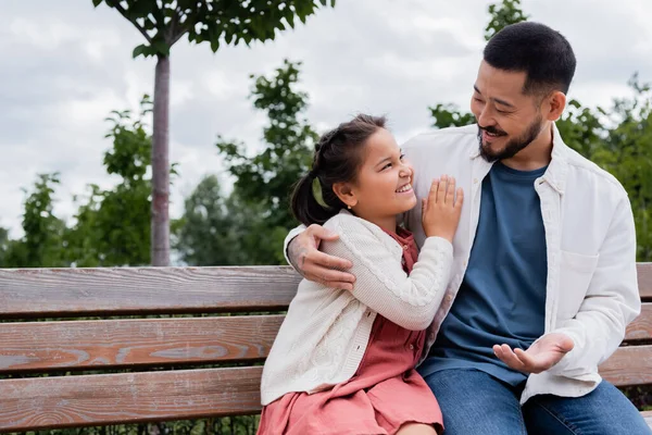 Smiling asian dad hugging and talking to daughter on bench in park - foto de stock