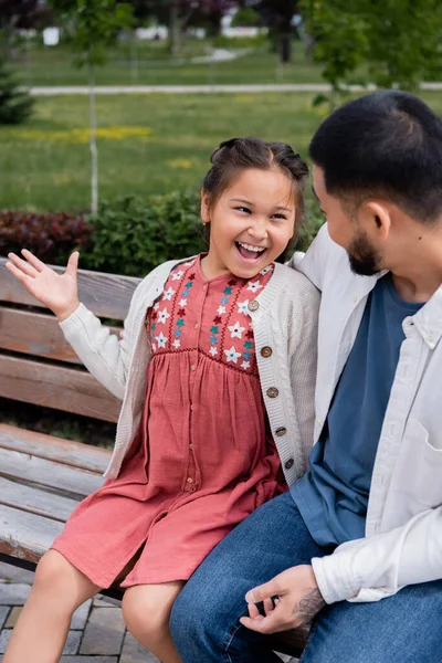 Excited asian girl talking to father on bench in park — Stock Photo