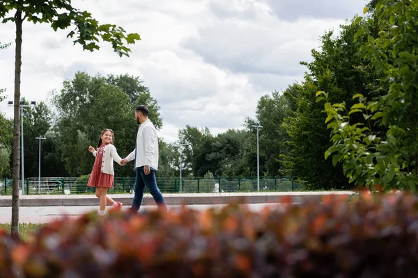 Улыбающаяся азиатская девушка, держащаяся за руку отца во время прогулки по летнему парку — стоковое фото