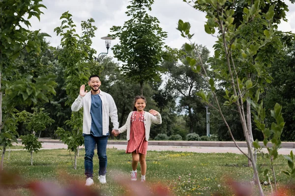 Smiling asian dad and daughter waving hands at camera in park — Stock Photo