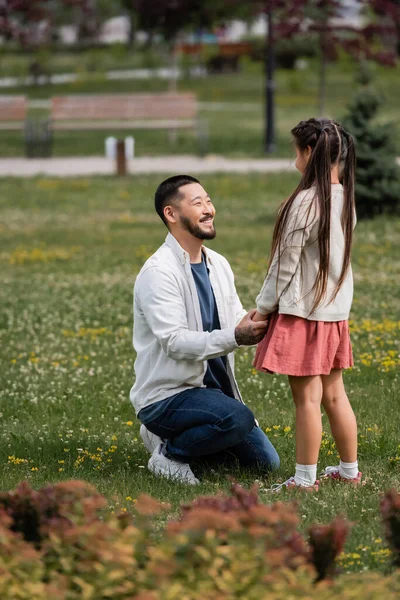 Happy asian parent holding hand of preteen daughter on meadow in park - foto de stock