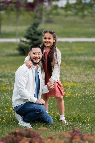 Cheerful asian kid hugging father on meadow with flowers in park — Photo de stock