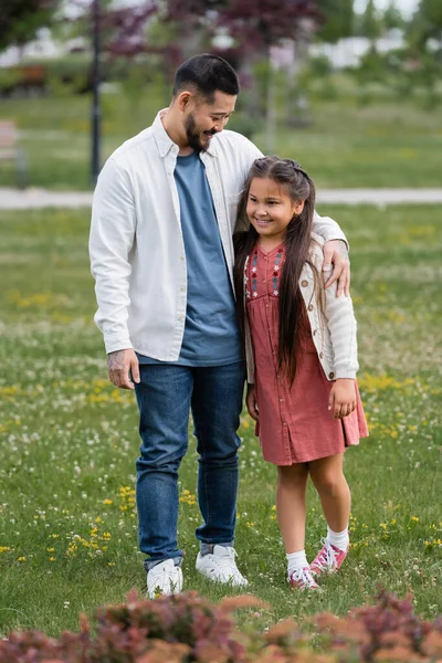 Positive asian dad hugging daughter on meadow with flowers in park — Stockfoto