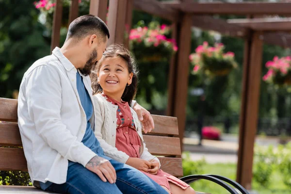 Cheerful asian girl looking at tattooed dad on bench in park — Stock Photo