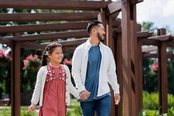 Asian father and preteen girl looking away while walking in park - foto de stock