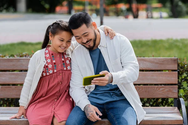 Позитивный азиатский папа держит смартфон рядом с дочерью на скамейке в парке — стоковое фото