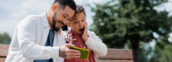 Азиатский мужчина держит сотовый телефон рядом изумленной дочери в парке, баннер — стоковое фото