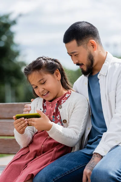 Asian parent hugging daughter with smartphone on bench in park — Stock Photo