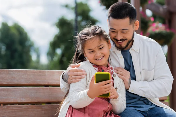 Усміхнений азіатський тато обіймає дочку зі смартфоном на лавці в парку — стокове фото
