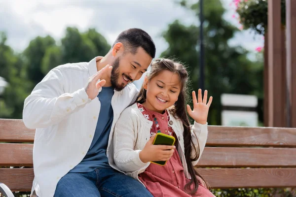 Positive asian father and daughter having video call on smartphone on bench in park — Fotografia de Stock