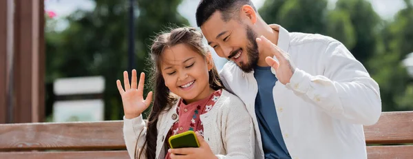 Азіатські батьки і дитина мають відеодзвінок на смартфоні на лавці літнього парку, банер. — стокове фото