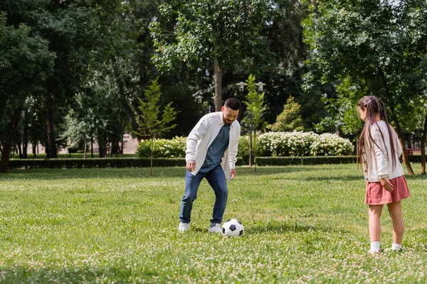 Усміхнений азіатський чоловік грає у футбол з дочкою на газоні в парку — стокове фото
