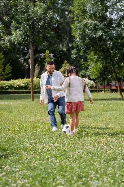 Positive asian dad playing soccer with daughter in park - foto de stock