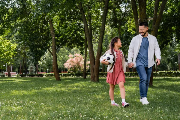 Smiling asian girl holding football and hand of father in park - foto de stock