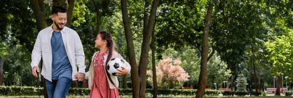 Улыбающийся азиатский папа держит за руку дочь-подростка с футболом в парке, баннер — стоковое фото