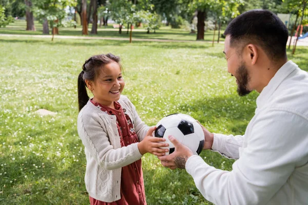Улыбающийся азиат держит футбольный мяч рядом с дочерью в летнем парке — стоковое фото