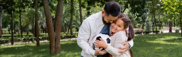 Cheerful asian dad holding football and hugging daughter in park, banner — Stockfoto