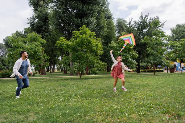Cheerful asian girl playing with colorful flying kite near dad running in park — Stock Photo