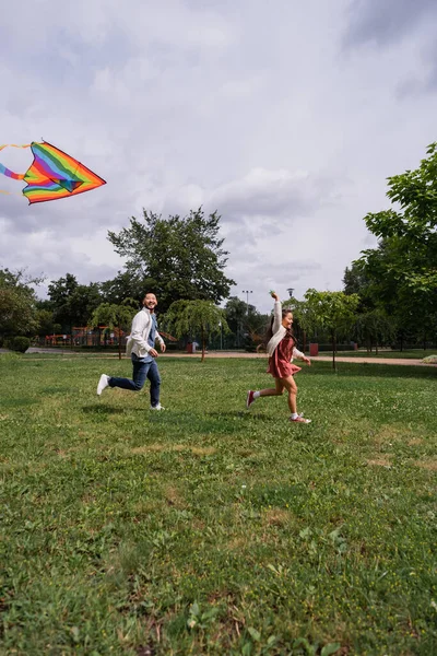 Positive asian man running near daughter with flying kite in park — Foto stock