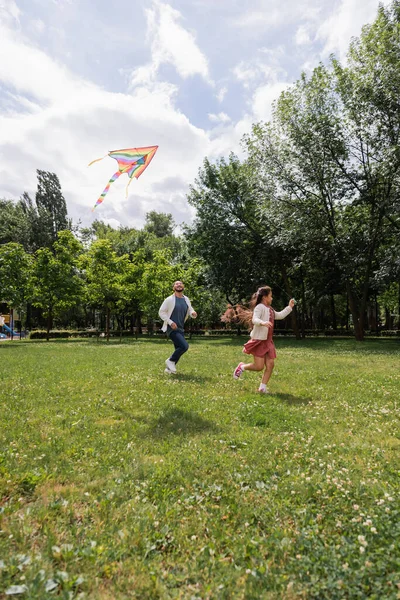 Позитивный азиатский родитель бегает рядом с дочерью с летящим воздушным змеем в парке — стоковое фото